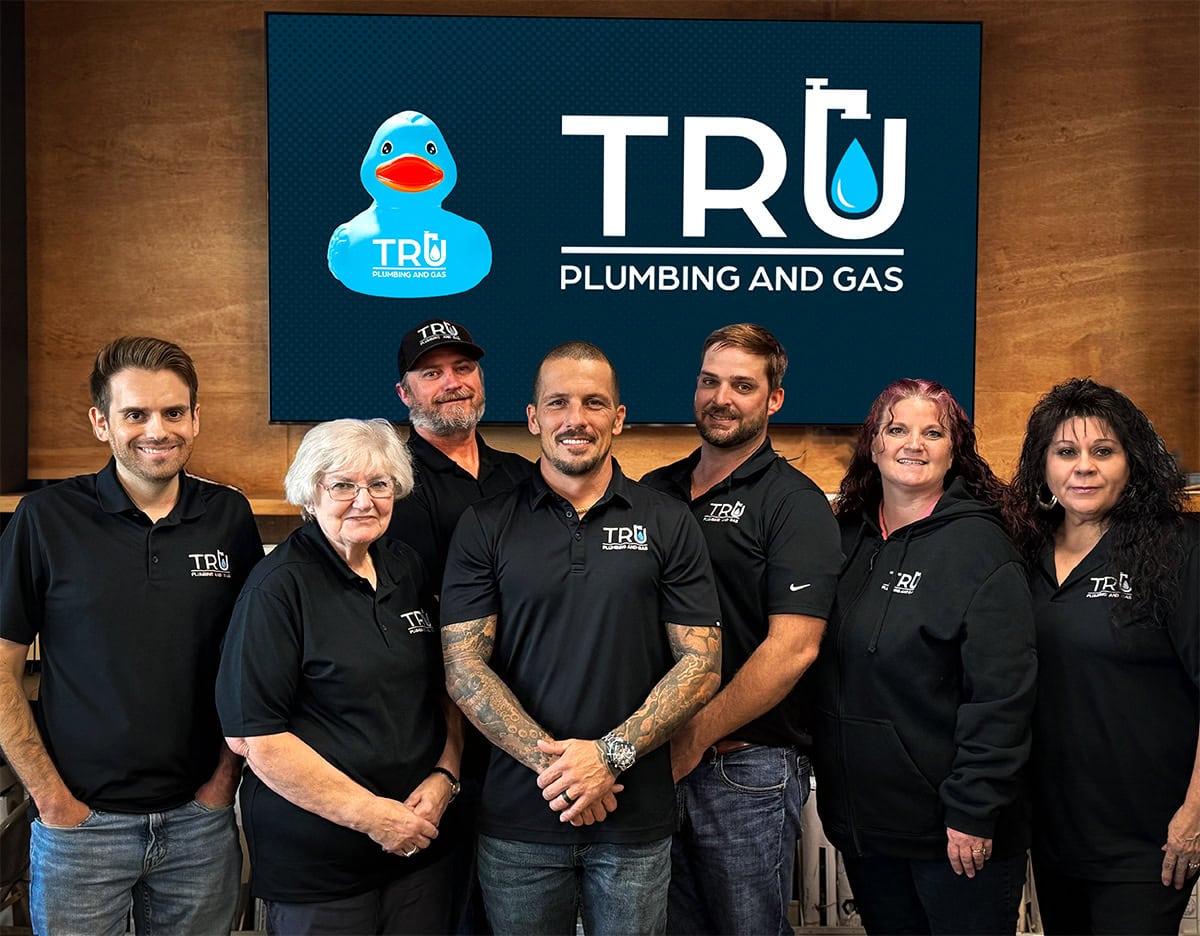 4 men and 2 women, smiling and standing in front of Tru Plumbing and Gas logo