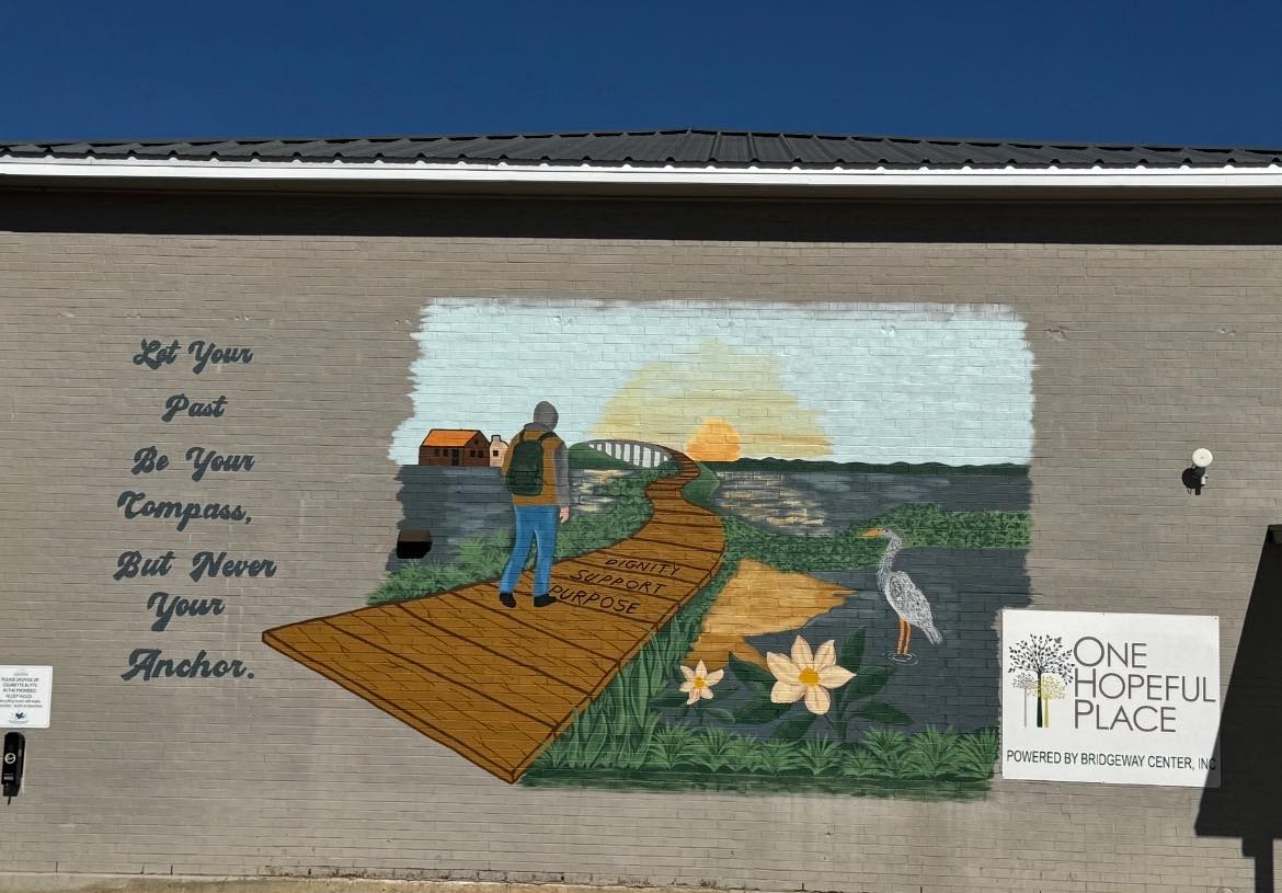 Painted mural on a brick wall of a man walking on a bridge with a sunrise in the distance. To the right is a sign for One Hopeful Place, and to the left are words reading "Let Your Past Be Your Compass, But Never Your Anchor"